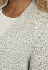 Light gray knitted cardigan with a textured pattern, featuring a v-neck and open front design, shown over a white top.