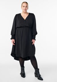 Mujer que lleva un vestido negro de manga larga con escote en V, pantys negros y botas negras hasta el tobillo, de pie contra un fondo blanco.