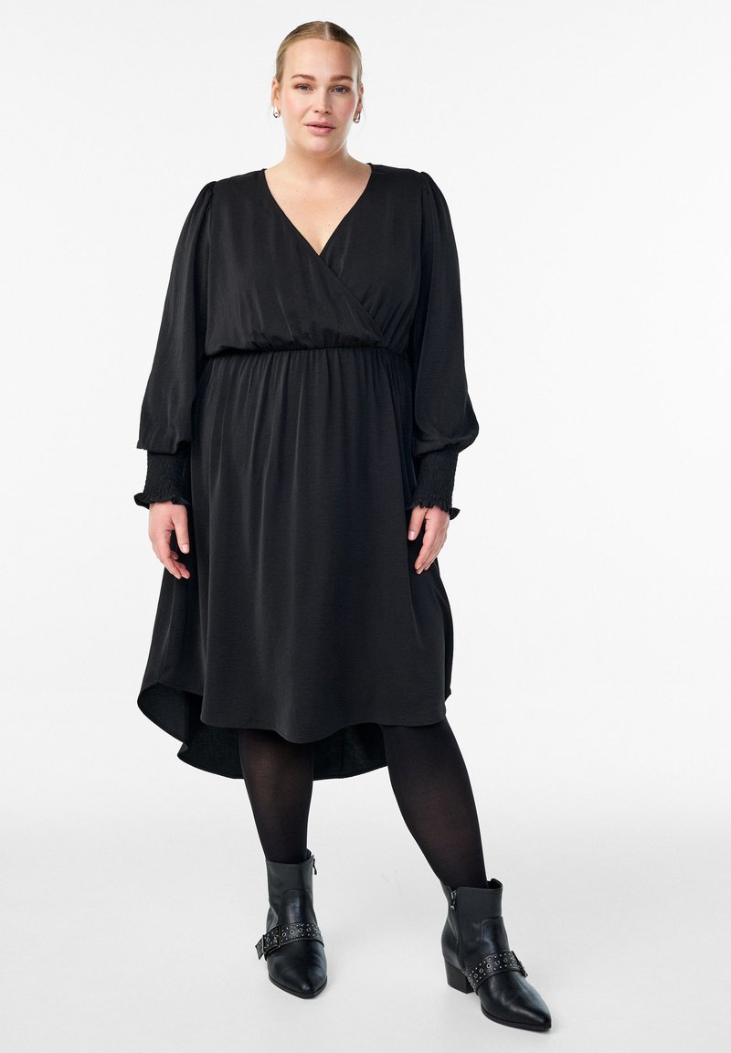 Mujer que lleva un vestido negro de manga larga con escote en V, pantys negros y botas negras hasta el tobillo, de pie contra un fondo blanco.