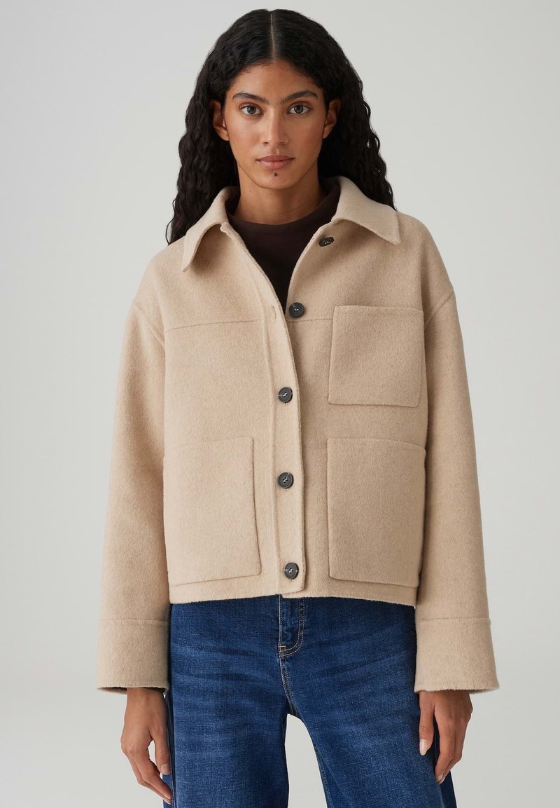 Veste beige en mélange de laine avec un col pointu, une fermeture à boutons sur le devant, deux grandes poches plaquées et des manches larges. Portée avec un jean bleu.