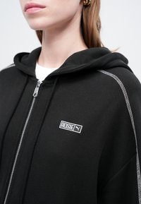 Žena nosi crnu duksericu na zip s bijelim šavom i "ROSIE" Puma logotipom na prsima, uparena s zlatnim kružnim naušnicama.