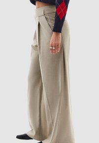 Béžové kalhoty s širokými nohavicemi vyrobené z texturované látky s sklady, s vysokým pasem a hladkým povrchem, stylizované s černými botami.