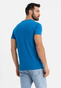 Modrá bavlněná tričko s krátkým rukávem a kulatým výstřihem, zobrazené na modelu zezadu, spojené se světle modrými džínami.