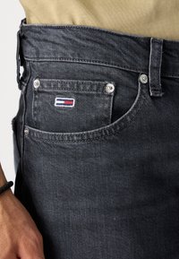 Jeans de mezclilla gris oscuro con un bolsillo frontal que presenta un pequeño logotipo bordado. La textura es suave, con sutiles detalles de costura.