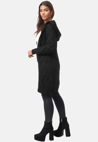 Cardigan lungo nero a maglia con cappuccio, caratterizzato da un motivo a trecce testurizzato. Abbinato a jeans skinny scuri e stivaletti con tacco.