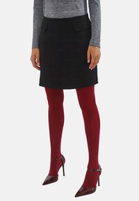 Czarna mini spódniczka z tweedu z dwiema przednimi kieszeniami, zestawiona z czerwonymi kryjącymi rajstopami i burgundowymi szpilkami. Widoczna jest także szara bluzka z długim rękawem.