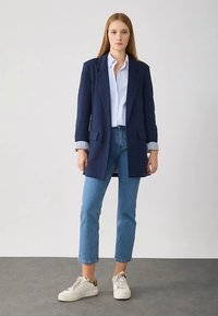 Laivastonsininen bleiseri, jossa on kaksi etutaskua, ryhdikäs muotoilu ja rento istuvuus, kerrostettuna vaaleansinisen paidan päälle ja yhdistetty vaaleansinisiin farkkuihin.