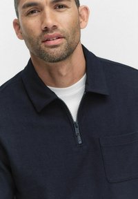 Marineblauw zip-up shirt met een kraag, met een textuurstof, een borstzak en een contrasterende witte binnenvoering. Ritsen en stiksels zichtbaar.