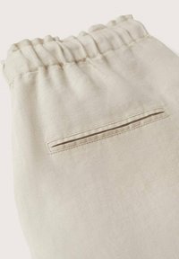 Pantalon en tissu beige avec ceinture élastique et une poche arrière horizontale fendue, montrant les détails de la texture et des coutures.