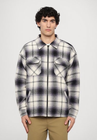 Plaid zip-up jacket in cream, black, and purple. Features two chest pockets and a collar. Soft, textured material with a relaxed fit.