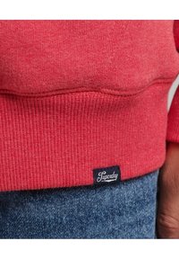 Close-up of red sweatshirt hem with black and white Superdry logo tag above blue denim jeans.