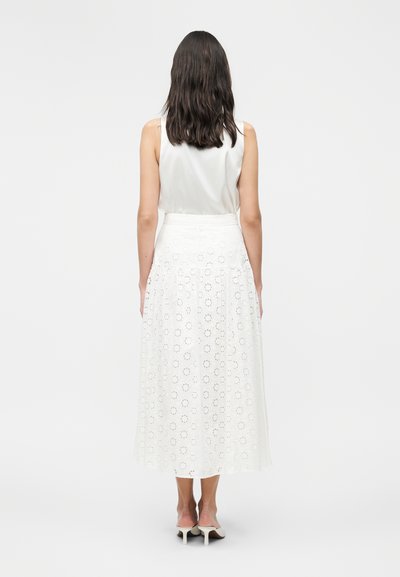 Γυναίκα με σκούρα μαλλιά φοράει λευκή αμάνικη μπλούζα, λευκή φούστα μέχρι τον αστράγαλο με λουλουδάτο δαντελένιο σχέδιο και λευκές γόβες, στέκεται με την πλάτη προς τα εμάς.