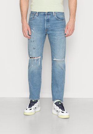 Mand iført lyseblå revnede jeans og hvide sneakers står mod en hvid baggrund med arme afslappet ned langs siderne.