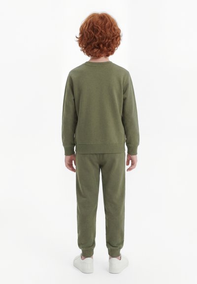 Dieťa s kučeravými červenými vlasmi oblečené v olivovo zelenej mikine a teplákoch, stojace otočené chrbtom, na bielom pozadí.