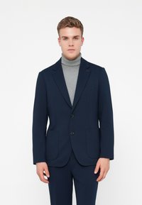 Jovem vestido com um fato azul marinho, com um casaco de dois botões e um suéter de gola alta cinza-claro, em pé contra um fundo branco.