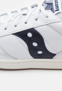 Zapatilla de cuero blanco con acentos de ante azul marino, que presenta tres cortes circulares, suela de goma texturizada y cordones blancos.