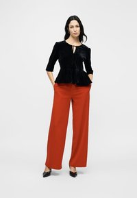 Black velvet peplum top with three-quarter sleeves and a tie detail; paired with wide-leg rust-colored trousers and black pumps.