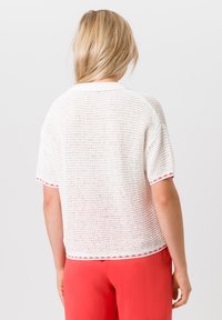 Weißes gestricktes kurzärmliges Oberteil mit strukturierter Musterung, verziert mit roten Akzenten an den Ärmeln und am Kragen. Kombiniert mit lebendigen korallenfarbenen Hosen.