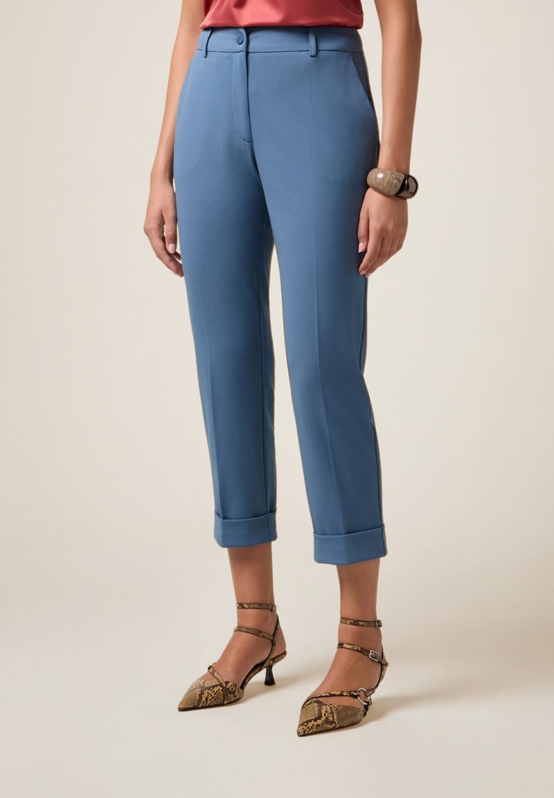 Donna che indossa pantaloni blu con risvolto, scarpe con tacco a punta in stampa serpente marrone con fasce alla caviglia, e un bracciale spesso sul polso sinistro.