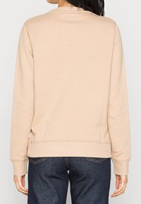 Sudadera de color beige claro con cuello redondeado, puños y dobladillo acanalados. Textura suave, ajuste relajado, diseño sencillo, llevada con jeans oscuros.