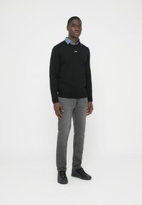 Juodas megztinis su V formos iškirpte ir baltu logotipu, dėvimas virš mėlynos marškinių apykaklės. Pilkos džinsinės kelnės ir juodi sportbačiai užbaigia įvaizdį.