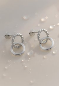 Silberne Ohrringe mit einem gedrehten Ringdesign, das einen glatten äußeren Kreis integriert. Dargestellt mit Tropfen auf einem sanften Hintergrund.
