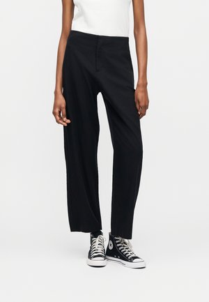 Άτομο που φοράει μαύρο παντελόνι με ψηλή μέση και ίσια γραμμή, λευκό αμάνικο τοπ και μαύρα ψηλά αθλητικά παπούτσια Converse, όρθιο σε λευκό φόντο.