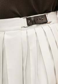 Κοντινό πλάνο κρεμ πλισέ φούστας με σκούρο καφέ δερμάτινη ζώνη στερεωμένη με μεταλλική αγκράφα, φορεμένη με σκούρο καφέ τοπ.