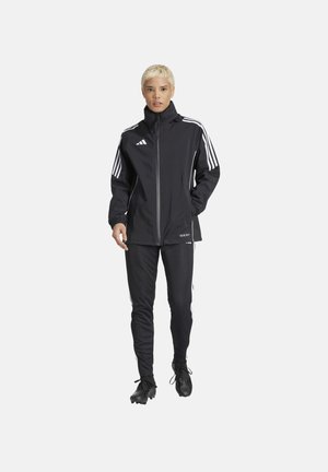 Schwarze Sportjacke mit weißen Streifen auf den Schultern, kombiniert mit schwarzen Hosen und schwarzen Sportschuhen. Verfügt über einen hohen Kragen und einen Reißverschluss.