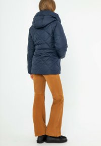 Námořnická prošívaná bunda s kapucí, s úzkým pasem a texturovaným povrchem, v kombinaci s hnědými kordovými zvonovými kalhotami a černými botami.