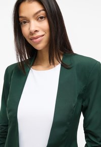 Vihreä bleiseri, jossa on sileä pinta ja rakenteellinen muotoilu, avoin etuosa, päällä yksinkertainen valkoinen paita, korostaen ammattimaista ilmettä.