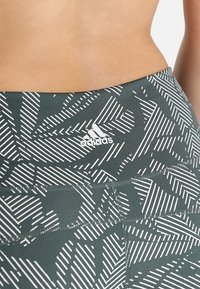 Legginsy sportowe o wysokim stanie z białymi geometrycznymi wzorami i logo Adidas z boku, pokazane na gołym brzuchu.