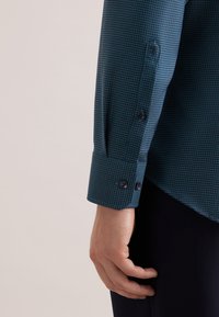 Donkergroenblauw shirt met een gestructureerde stof, een nauwsluitende kraag en manchetten met knopen, voorzien van twee knopen op de manchet voor aanpassing.