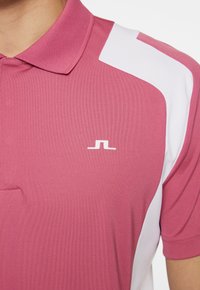 Rosa polotröja med vita axelaccenter, en klassisk krage och en liten vit logotyp. Tygmaterialet verkar vara slätt och stretchigt.