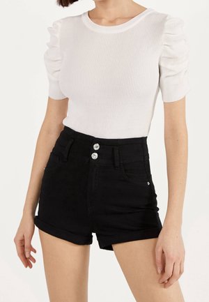 Kvinde iført en hvid ribbet kortærmet top med puffede skuldre og højtaljede sorte shorts med dobbeltknapper.
