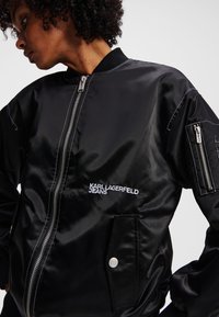 Juodas satino bomberio tipo švarkas su užtrauktukais, šoniniais ir rankovės kišenėmis, o balta siuvinėta užrašas „KARL LAGERFELD JEANS“. Per didelis kirpimas.