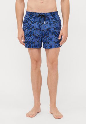 Homme portant un short de bain bleu avec un cordon noir et un grand motif répété du logo "DSQUARED2", debout pieds nus sur fond blanc.