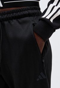 Άτομο που φοράει μαύρο παντελόνι Adidas με ελαστική μέση και τσέπη με φερμουάρ, χέρι μέσα στην τσέπη, ορατό λογότυπο Adidas κοντά στο γόνατο.