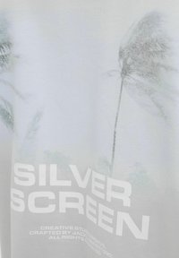 Balts T-krekls ar izbalējušu palmu attēlu un izteiksmīgu tekstu "SILVER SCREEN" lielā baltā sans-serif fontā. Gluda kokvilnas tekstūra.