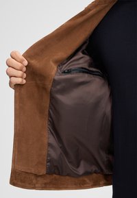 Binnenkant van een bruine suede jas, met een gladde donkerbruine voering en een ritszak voor veilige opslag.