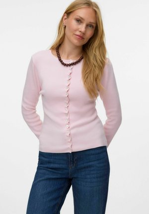 Femme aux longs cheveux blonds portant un pull rose à boutons, un collier foncé et un jean bleu, debout les mains dans le dos.