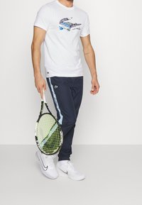 White t-shirt with colorful Lacoste crocodile logo, paired with navy track pants featuring light blue side stripes, and white Nike sneakers.