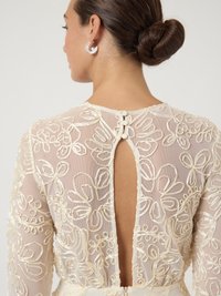 Διάφανο, κεντητό φόρεμα με floral σχέδια σε ελεφαντόδοντο, με χαμηλή πλάτη και λεπτομέρειες από κουμπιά, συνδυασμένο με ασημένια κυκλικά σκουλαρίκια.