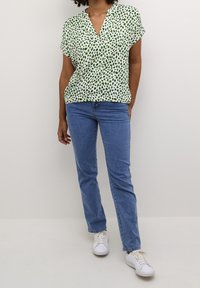 Green and white short-sleeve blouse with a v-neck, featuring a scatter print design. Paired with light blue jeans and white sneakers.