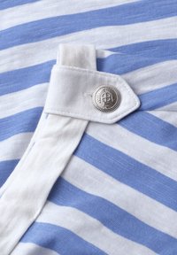 Bouton argenté avec emblème d'arbre sur un onglet en tissu blanc au-dessus d'un tissu en coton rayé bleu et blanc.