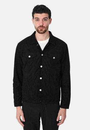 Uomo che indossa una giacca nera con bottoni e tasche sul petto sopra una camicia bianca, in piedi davanti a uno sfondo bianco semplice.