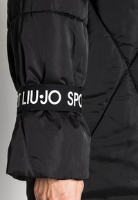 Manga de chaqueta acolchada negra con un diseño texturizado y acolchado. Puño elástico con una banda blanca contrastante que dice "SPORT LIU·JO."