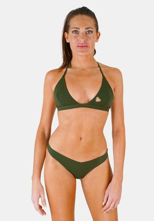 Femme aux longs cheveux foncés portant un bikini vert avec un petit emblème en forme de cœur sur le haut, debout devant un fond clair uni.