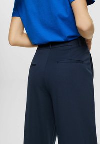 Person som bär skräddarsydda navyblå höga byxor och en instoppad ljusblå kortärmad skjorta mot en neutral bakgrund.