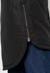 Veste noire avec une fermeture éclair argentée verticale partiellement ouverte sur un jean en denim bleu, montrant un détail d'ourlet incurvé sur le côté.